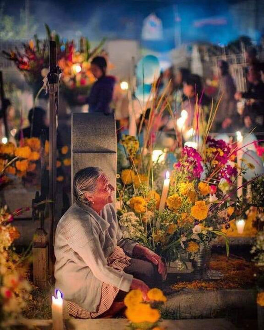 Elderly woman sits by a tomb, adorned with candles and flowers